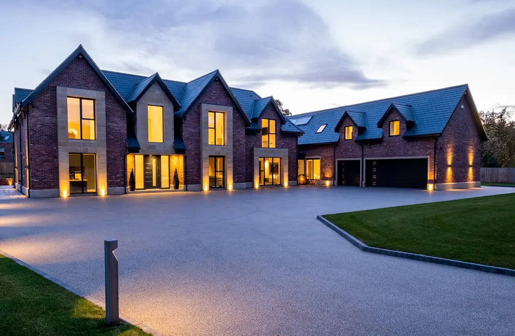 Exterior of a large home lit at dusk, with uplighting on the brickwork and a lit driveway